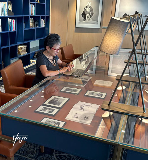 A person working on a laptop at a stylish desk with framed photographs displayed underneath a glass surface in an elegantly designed library or study area.