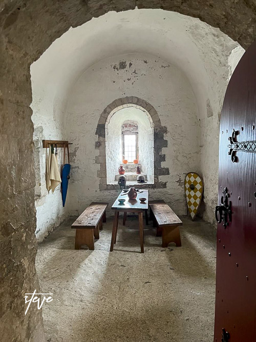 Interior of a medieval room with an arched ceiling, featuring a wooden table and benches, decorative pottery, and shields on the walls.
