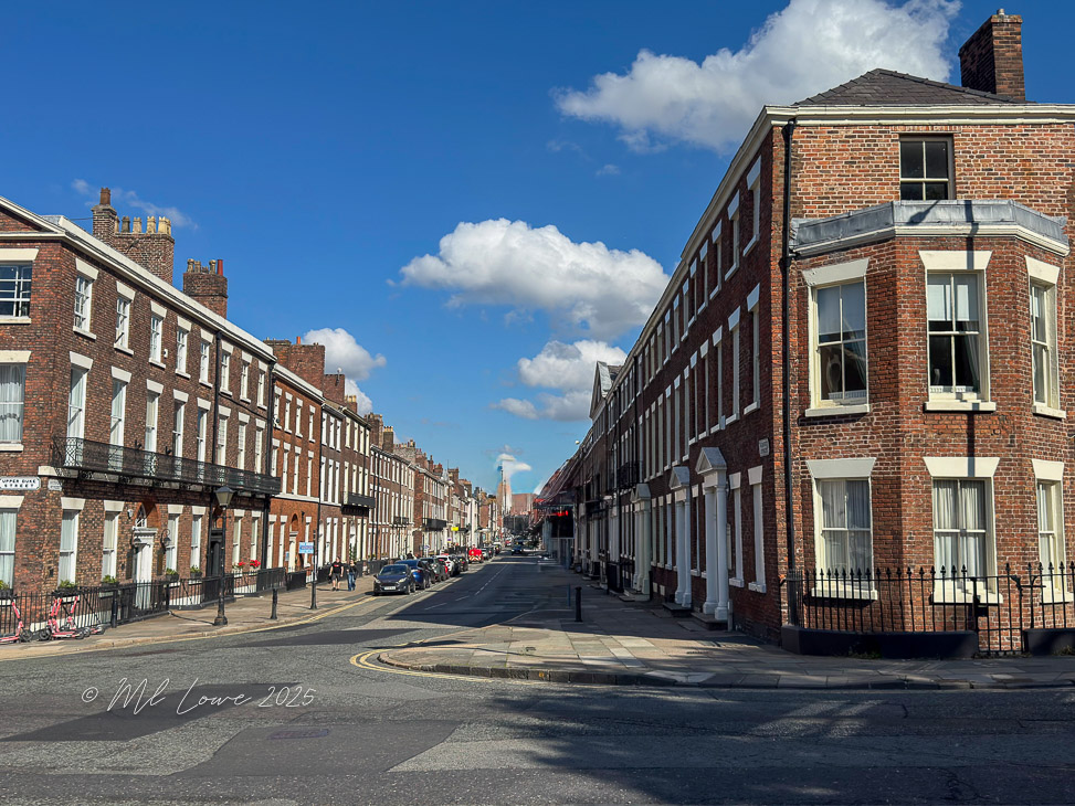 A street view of brick buildings along a road in a city, with blue skies and fluffy clouds in the background.