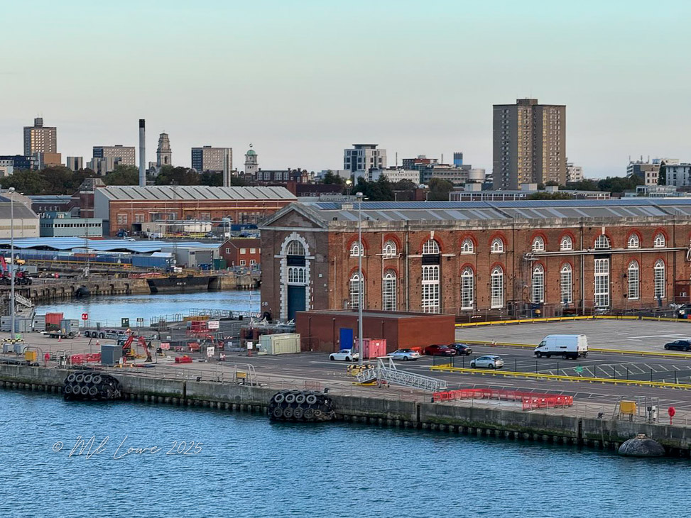 A view of a waterfront area with a red-brick building and modern structures in the background. The scene includes parked vehicles and a calm water surface.