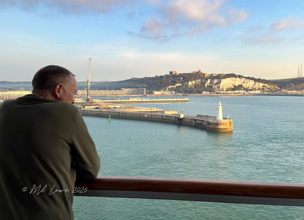 A person gazing at the view from a cruise ship balcony, overlooking a harbor with a lighthouse and cliffs in the background.