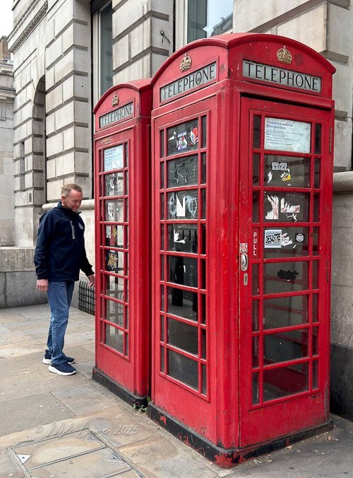 A man stands in front of two classic red telephone boxes in London, examining the interior while surrounded by historic architecture.