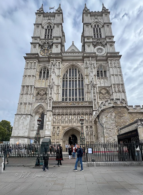 The front view of Westminster Abbey, showcasing its iconic Gothic architecture, with visitors gathering at the entrance and cloudy skies above.
