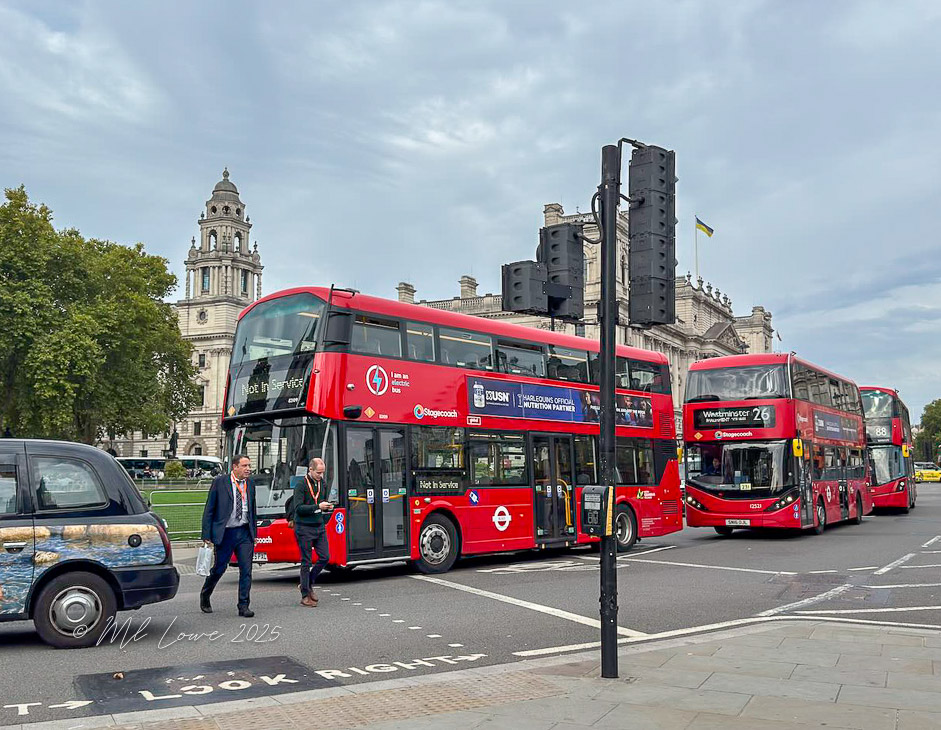 Busy street scene in London featuring iconic red double-decker buses and pedestrians crossing at a traffic signal.