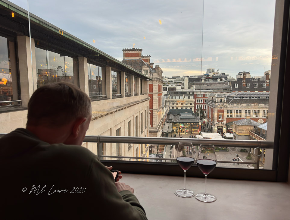 A person sitting by a window in a London café, looking at their phone, with two glasses of red wine on the table and a view of the bustling streets below.