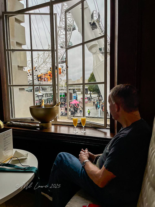 A person sitting at a window in a cafe, gazing at the London Eye outside, with two drinks on the table.