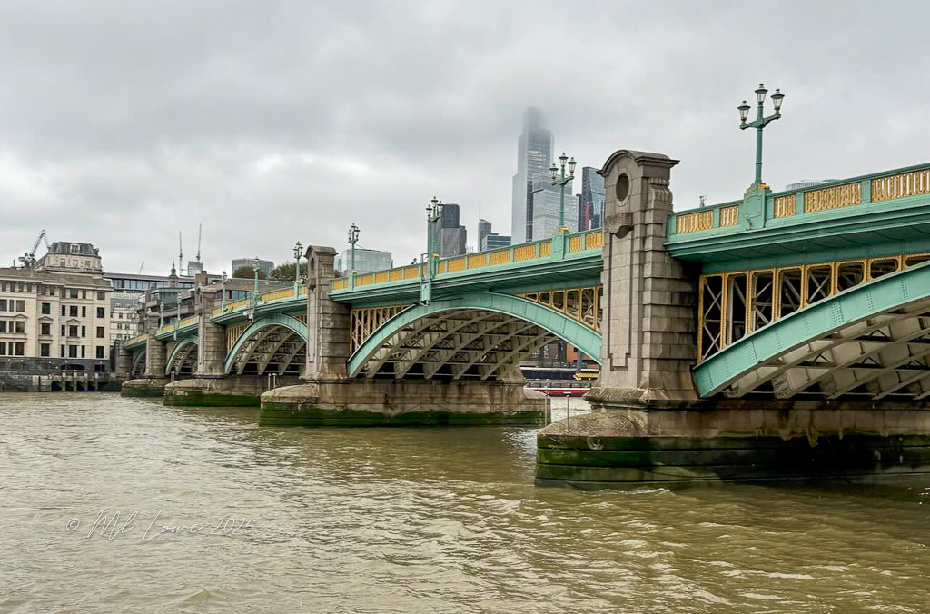 View of a green and gold bridge over the River Thames in London, with modern skyscrapers in the background on a cloudy day.