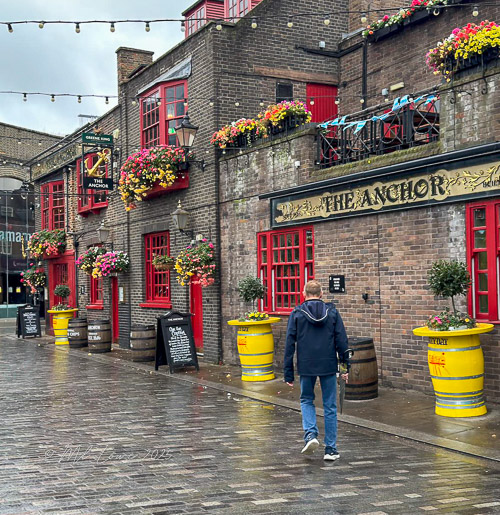 A person walking past a pub called 'The Anchor', featuring red accents and vibrant flower decorations in London.