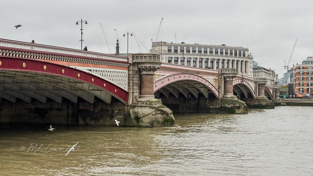 View of a bridge over the River Thames in London, showcasing its arch design and architectural details, surrounded by city buildings and a cloudy sky.