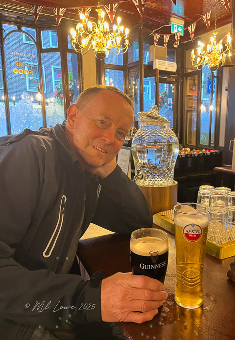 A man sitting in a pub, smiling while resting his head on his hand. He has a glass of Guinness beer in front of him, alongside a glass of Amstel beer, with warm lighting and decorative elements in the background.