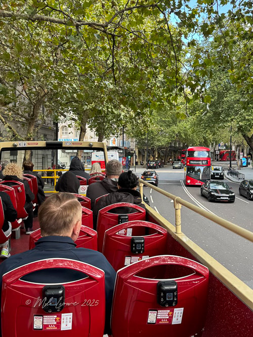 View from the upper deck of a red double-decker bus in London, showing passengers seated and busy city streets with traffic.