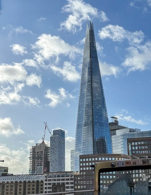 The Shard, a modern glass skyscraper in London, towers against a blue sky with scattered clouds, surrounded by other buildings under construction.