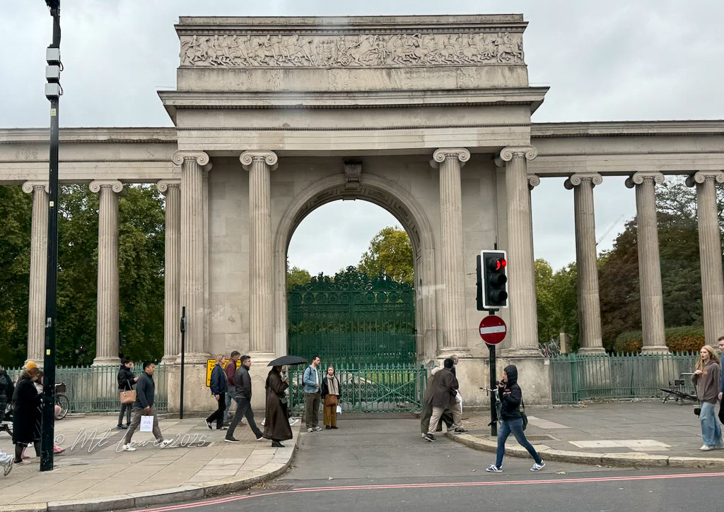 A historic archway with columns, leading to a green gate, surrounded by pedestrians crossing the street on a cloudy day in London.