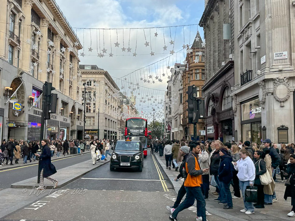 A busy London street scene with pedestrians, a red double-decker bus, and decorative lights hanging overhead.