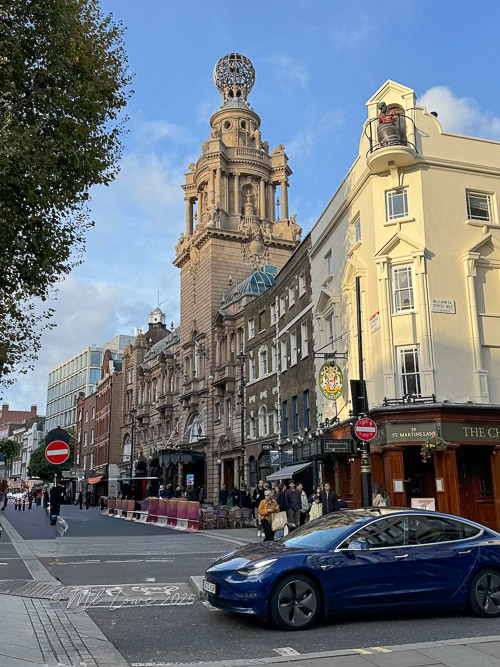Street view showcasing a historic building with a distinctive tower in London, featuring a mix of classic architecture, modern shops, and a blue car parked in the foreground.