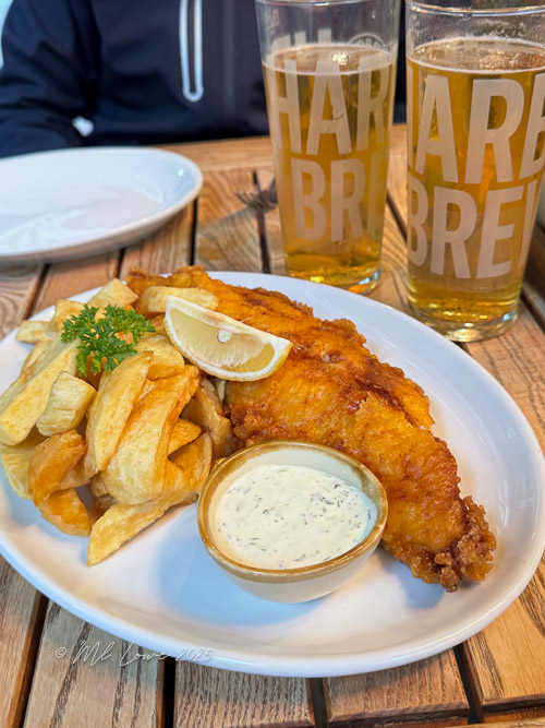 A plate of fish and chips served with a lemon wedge and tartar sauce, accompanied by two glasses of beer, on a wooden table.