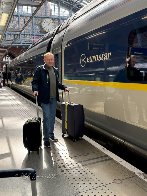 A man standing on a train platform beside a Eurostar train, holding two suitcases, with a clock visible in the background.