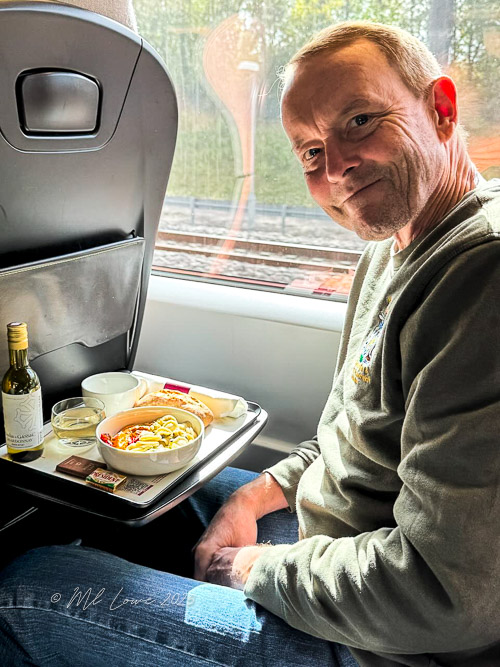 A person sitting on a train, smiling while holding a tray with a meal that includes pasta, a small bottle of wine, and a few snacks, with a blurred background of the train window.