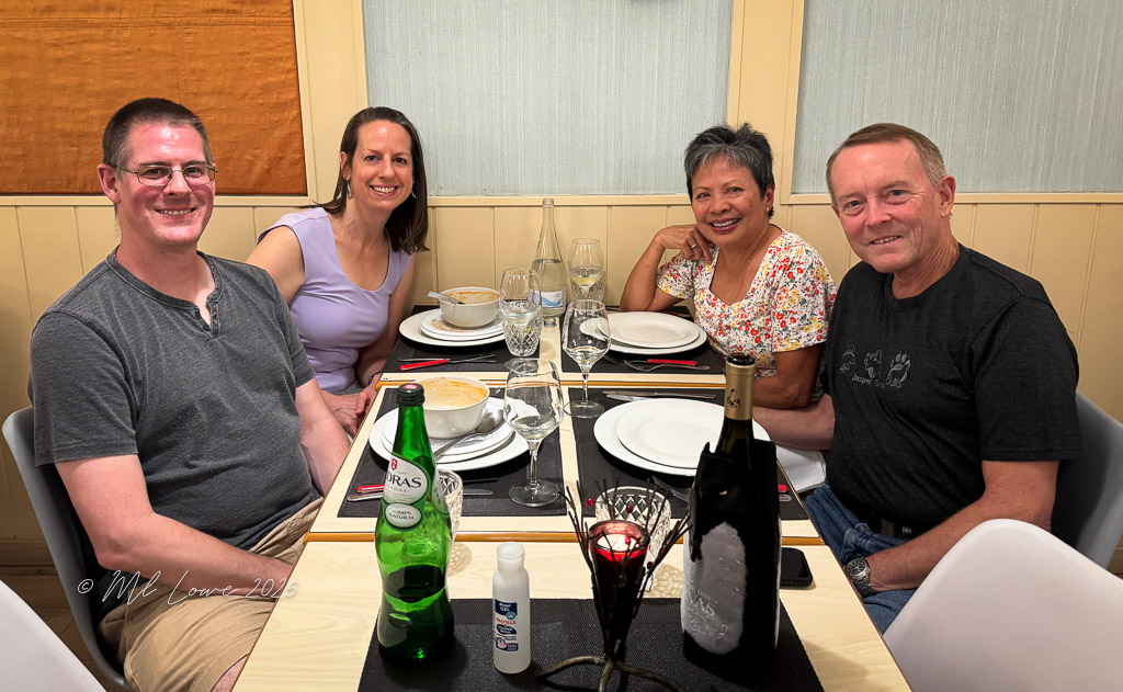 Four friends sitting at a dining table, enjoying a meal together. The table is set with plates, glasses, and bottles of water and wine.