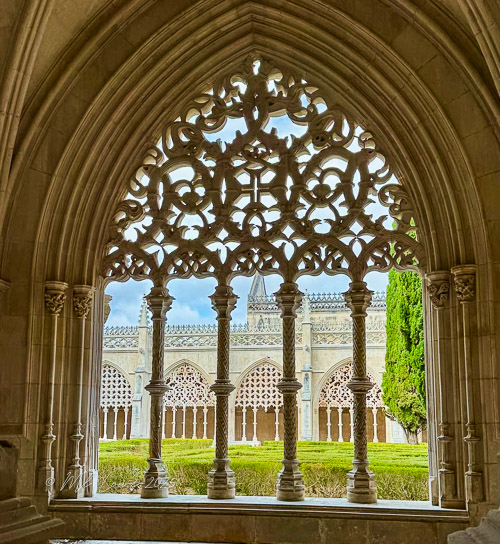 Intricate stone window framing a view of a lush garden and architectural details at Batalha Monastery in Portugal.