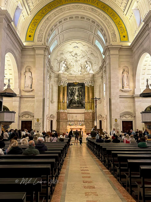 Interior view of the Sanctuary of Our Lady of Fátima, showing a crowd of visitors in a spacious, ornate church with a high ceiling and religious artwork.
