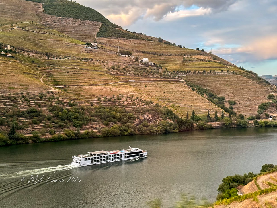 A scenic view of the Douro River with a river cruise ship navigating through the terraced vineyards and hilly landscape in Portugal.