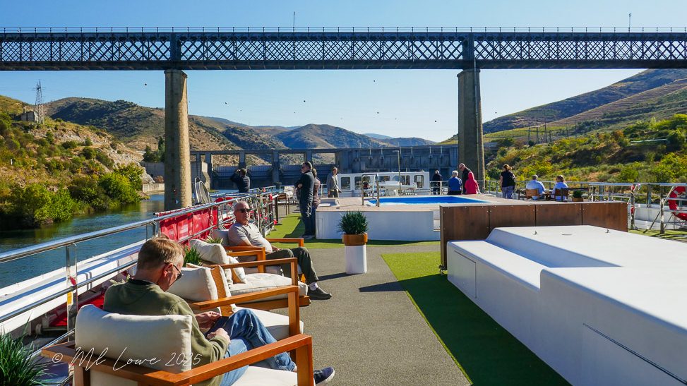 View of a Viking river cruise ship's deck with passengers relaxing, set against a backdrop of a bridge and rolling hills along the Douro River.