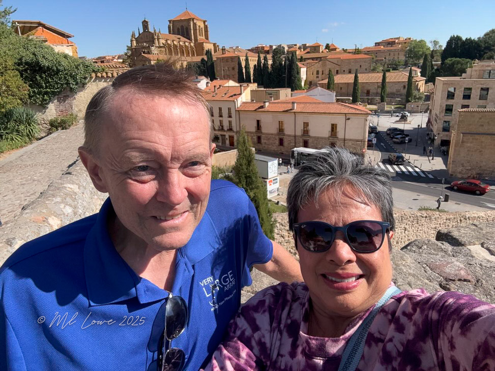 A couple poses for a selfie on a historic wall with a view of a picturesque town in the background featuring red-roofed buildings and greenery.