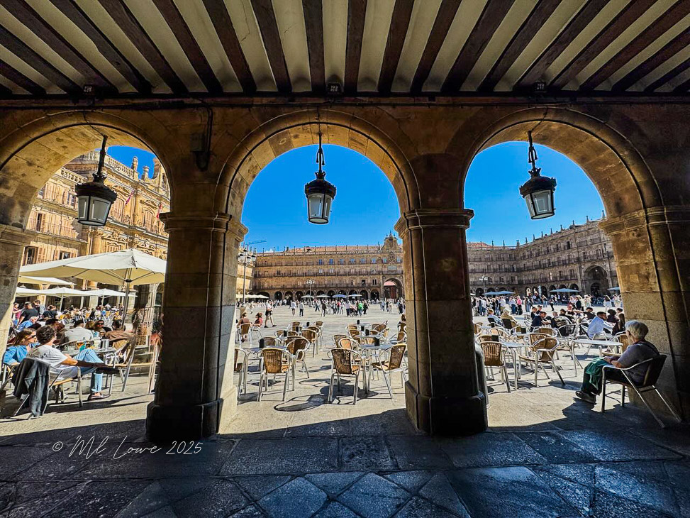 View from an arcade showcasing the vibrant Plaza Mayor in Salamanca, Spain, filled with people dining at outdoor cafes and enjoying the sunny day.