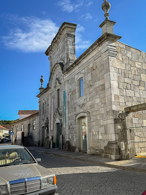 A historic stone building with intricate architectural details, set against a clear blue sky, alongside a vintage car parked on a cobblestone street.