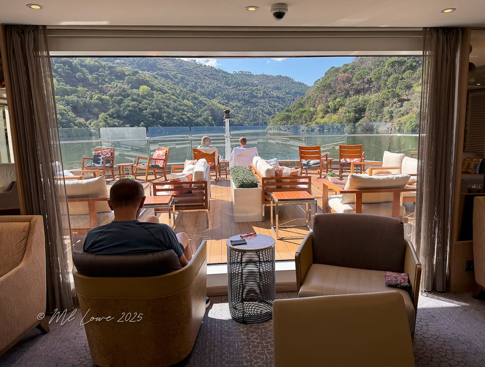 Interior view of a river cruise ship lounge with comfortable seating and large windows showcasing lush green mountains and calm waters outside.