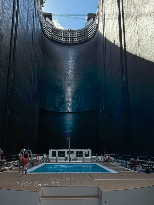 View of a river lock with a swimming pool visible on the deck of a river cruise ship, surrounded by steep, dark walls of the lock.