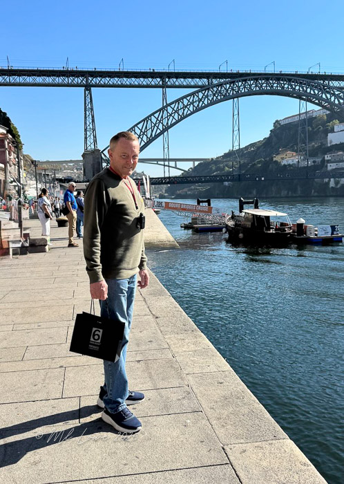 A person standing along a waterfront in Porto, Portugal, with a bridge in the background and boats moored nearby.