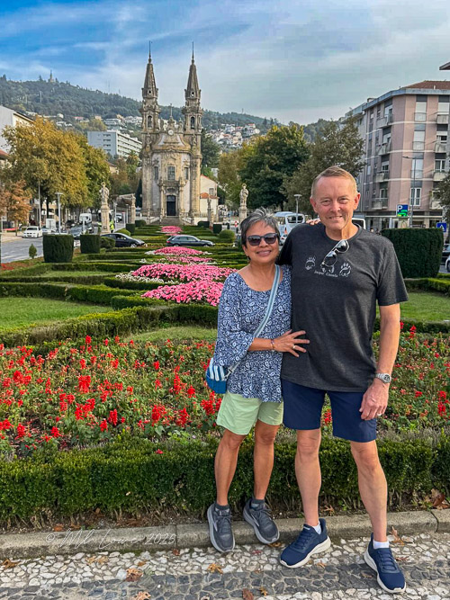 A couple stands together in a garden filled with flowers, with a church and trees in the background.