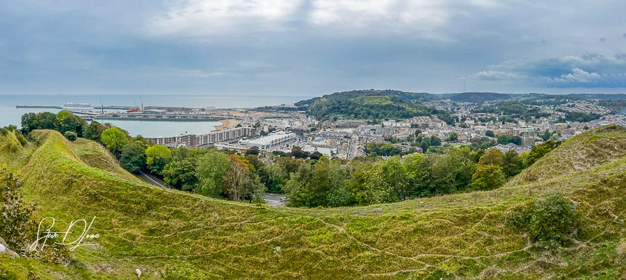 Panoramic view of the coastal city of Dover, England, showcasing the harbor, residential buildings, and rolling green hills in the foreground under a cloudy sky.