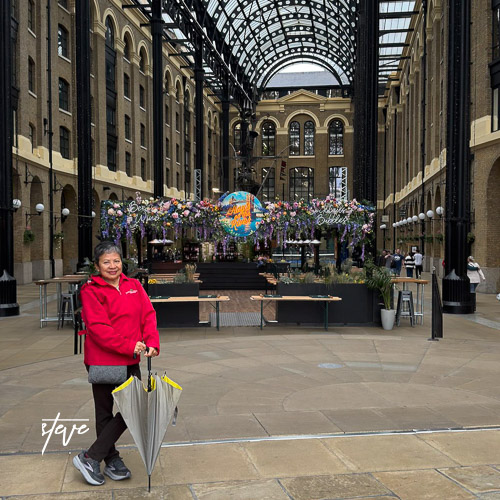 A person in a red jacket holding an umbrella stands in a spacious indoor market area featuring floral decorations and large windows.