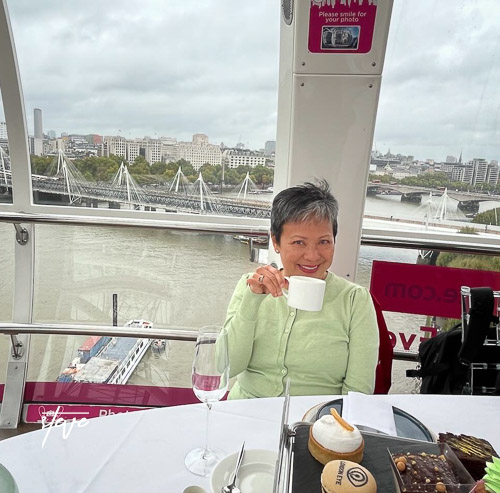 A woman enjoying afternoon tea while seated in a pod of the London Eye, with a view of the River Thames and cityscape in the background.