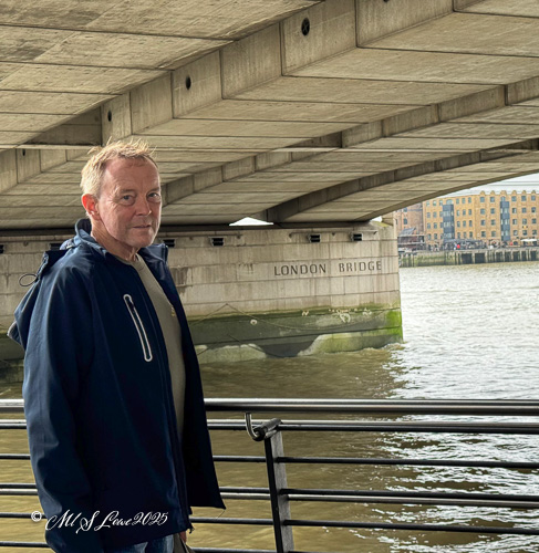 A person standing on a railing next to the River Thames with the London Bridge visible in the background, under a concrete arch.