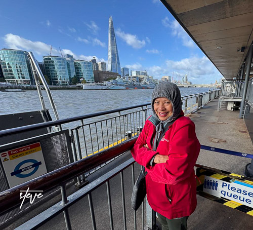 A person wearing a red jacket stands by the River Thames with the Shard visible in the background, showcasing the skyline of London.