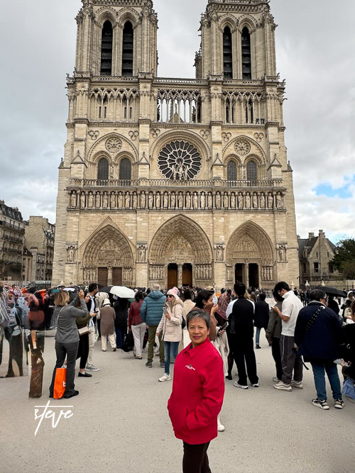 A woman in a pink jacket stands in front of a crowd outside the Notre-Dame Cathedral, showcasing its Gothic architecture with tall towers and intricate details.