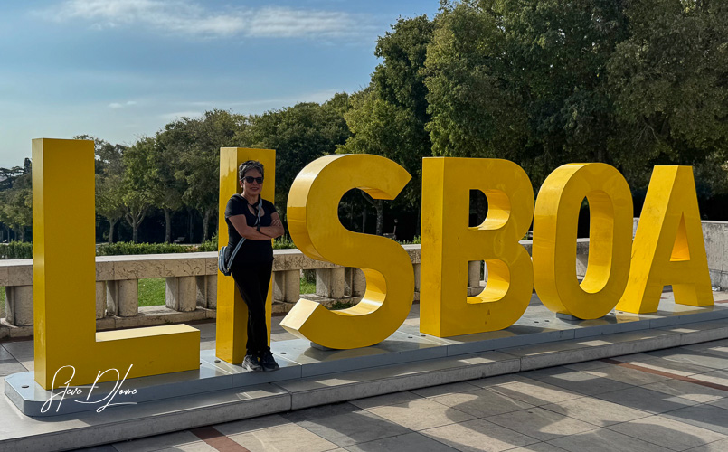 A person leaning against large yellow letters spelling 'LISBOA' in a park setting, surrounded by trees.