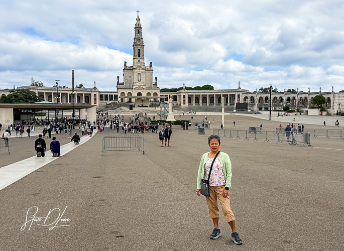 A woman stands in the foreground of a large open area at the Sanctuary of Our Lady of Fátima, with a prominent church building and numerous visitors in the background under a cloudy sky.