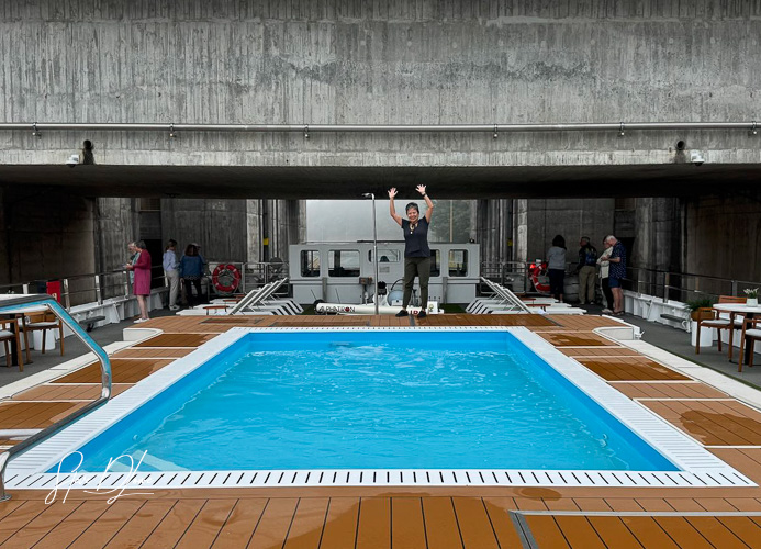 A person standing by a swimming pool on the Viking Osfrid river cruise ship, with water and wooden deck visible, while others are seen in the background.