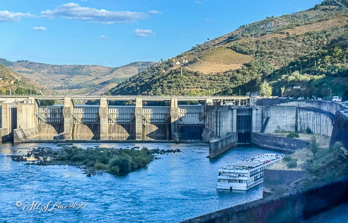A view of a dam on the Douro River, with a cruise ship navigating nearby, surrounded by green hills and a blue sky.