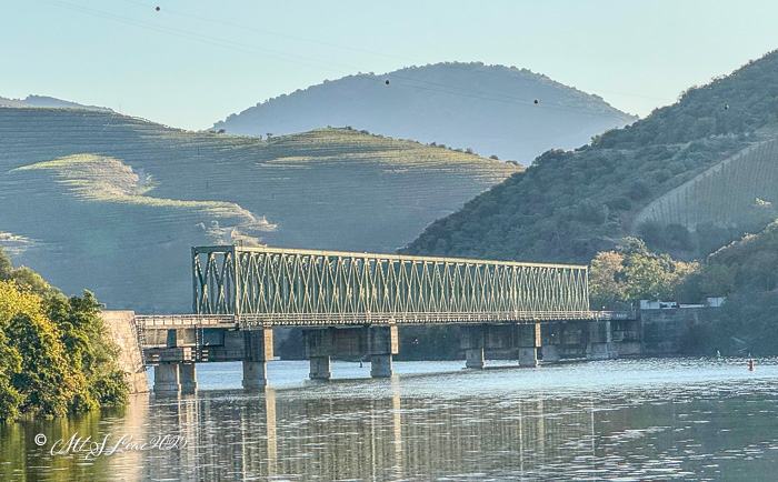 A green steel bridge spanning a calm river with rolling hills and terraced vineyards in the background, reflecting the warm morning light.