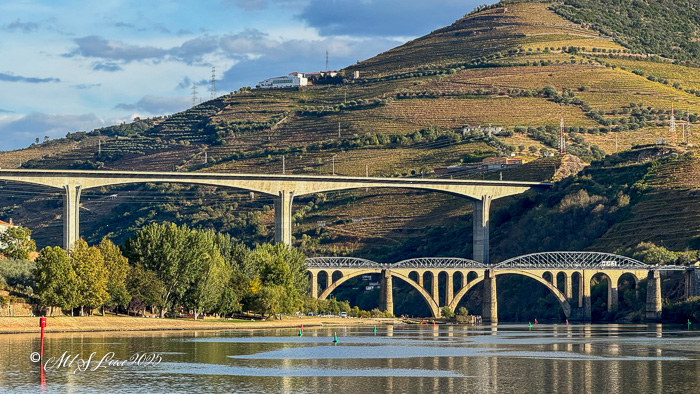 A scenic view of the Douro River featuring two distinct bridges: a modern arch bridge above and an older stone bridge below, surrounded by terraced vineyards on the hillside.