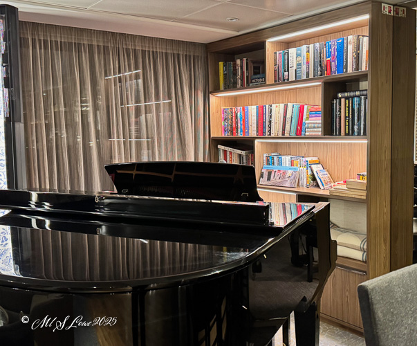 Interior view of a cozy room on a river cruise ship, featuring a black piano in the foreground and bookshelves filled with books in the background.