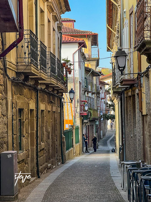 Narrow cobblestone street lined with historic buildings and balconies, with two people talking in the distance under a clear blue sky.