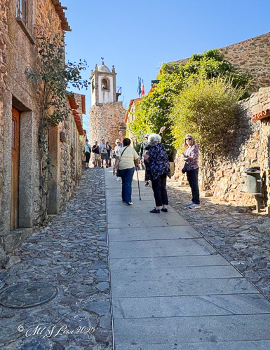 A narrow cobblestone street in a historic village with tourists walking up the path, featuring rustic stone buildings and a bell tower in the background.