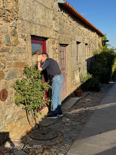 A person peering through a window of a stone building, surrounded by greenery and a cobblestone pathway.
